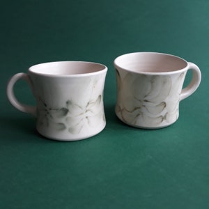Peut inclure: Deux tasses en céramique blanche avec un motif floral dans des tons de gris. Les tasses ont une forme arrondie et une poignée sur le côté.
