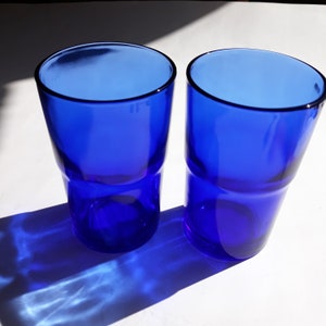 May include: Two cobalt blue glass tumblers, one slightly out of focus, on a white surface. The glasses are empty and have a rounded shape.