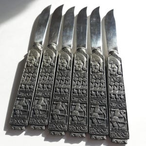 May include: Set of six silver-tone butter knives with intricate engraved designs. Each knife features a repeating pattern of a ship and the text "KIEER SAVER" and "AVD OTTER".