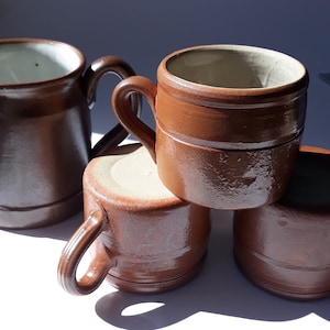 May include: Four brown ceramic mugs with handles. The mugs are stacked on top of each other. The mugs have a rustic, handmade look.