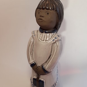 May include: A ceramic figurine of a girl with brown hair, wearing a white dress with blue trim. She has a blue bow in her hair and is holding a brown object in her hands.