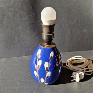 May include: A blue ceramic table lamp with a white and gold floral design. The lamp has a white light bulb and a white cord with a plug.