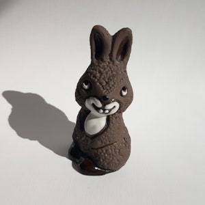 Könnte beinhalten: Eine braune Keramik-Hasenfigur mit weißen Details. Der Hase hat ein breites Lächeln und sitzt mit eingezogenen Pfoten.