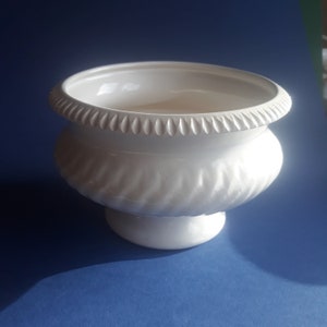 Op de afbeelding: Witte keramische plantenpot met een geribbeld ontwerp en een decoratieve rand. De pot heeft een brede basis en een rond lichaam.