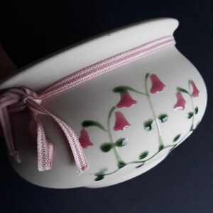 May include: A cream ceramic plant pot with a pink and white striped ribbon tied around the rim. The pot is decorated with pink bell-shaped flowers and green stems. Suitable for indoor or outdoor use, it is a decorative item.