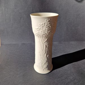 May include: White ceramic vase with a floral design. The vase is tall and slender with a wide mouth.