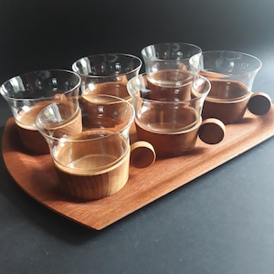 May include: A set of six clear glass cups with wooden holders and handles, presented on a matching wooden serving tray. The cups have a rounded shape, and the wooden elements are a warm brown color. The tray has a curved edge.