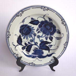 Op de afbeelding: Witte keramische plaat met een blauw bloemmotief. De plaat heeft twee grote bloemen, bladeren en decoratieve randelementen. De plaat wordt gepresenteerd op een zwarte standaard.
