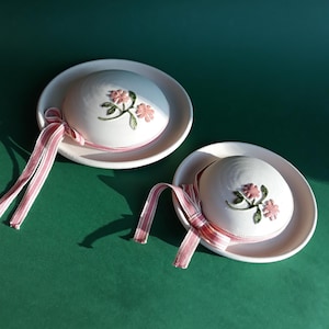 May include: Two white ceramic hat-shaped trinket boxes with pink and green floral designs. Each box has a pink and white striped ribbon.