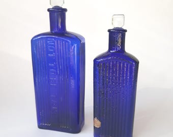 May include: Two antique cobalt blue glass bottles with clear stoppers. The larger bottle has the text "NOT TO BE TAKEN" embossed on the front. Both bottles have vertical ribbed detailing and are displayed against a white background.