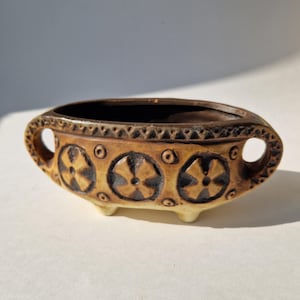 May include: A small, oval ceramic bowl with a brown interior and exterior. The exterior has a tan base with brown floral designs and a decorative border. The bowl has two handles and three small feet.