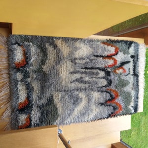 May include: A gray, black, and orange rug with a shaggy texture and a fringe border. The rug has an abstract design with a variety of shapes and colors.