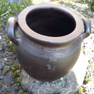 Könnte beinhalten: Ein brauner Keramiktopf mit zwei Henkeln. Der Topf hat das Wort "HOGANAS" und die Zahlen "2L" auf der Seite eingraviert. Der Topf steht auf einem Stein.