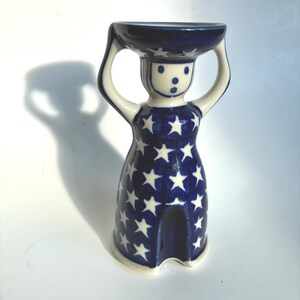 Pode incluir: Um suporte para ovo em cerâmica com a forma de uma figura estilizada com os braços levantados para apoiar uma taça. A figura é decorada com um vidrado azul escuro com um padrão repetitivo de estrelas brancas. O rosto tem pontos azuis.