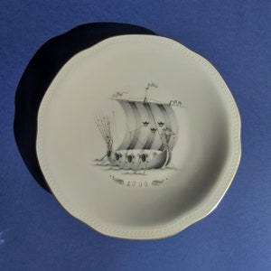 May include: A white ceramic plate with a gold rim features a black and white illustration of a Viking ship with sails. The ship has a dragon head at the bow and the number "1000" is printed below the ship.