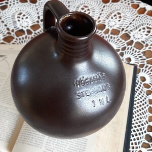 Peut inclure: Une cruche en céramique brune avec une anse. La cruche porte l'inscription "Hogan's Stein Gods 1 1/2 L" imprimée sur le côté.