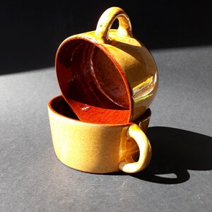 Puede incluir: Dos tazas de cerámica, una apilada sobre la otra. Las tazas son de color canela moteado en el exterior con un interior marrón rojizo intenso. Cada taza tiene un asa redondeada. Las tazas están sobre un fondo oscuro.