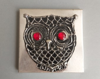May include: A silver metal wall art piece featuring a black owl with red glass eyes. The owl is textured and has a detailed design.