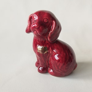 Op de afbeelding: Een klein, rood keramiek beeldje van een hond die zit met zijn hoofd gekanteld. Het beeldje heeft een glanzende afwerking en is gemaakt door Guldkråken Hjo Zweden.