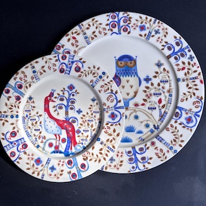 Peut inclure: Deux assiettes en céramique blanche ornées de motifs d'art populaire représentant des arbres, des fleurs et des oiseaux stylisés. La grande assiette présente un hibou bleu, tandis que la petite montre un oiseau rouge et bleu, avec du feuillage brun.