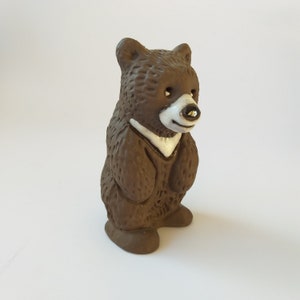 May include: A small brown ceramic bear figurine with white accents around the eyes and mouth. The bear is standing upright with its paws together.