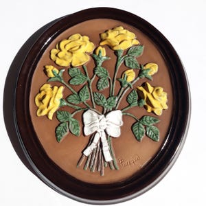 May include: Oval wall plaque featuring a bouquet of yellow roses with green leaves and buds, tied with a white bow. The relief sculpture is set against a brown background and framed in a dark brown oval.