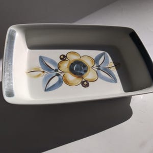 May include: Rectangular ceramic serving dish with a white base and a floral design. The flower has yellow and blue petals with a dark blue centre. The dish has a grey border on the sides.