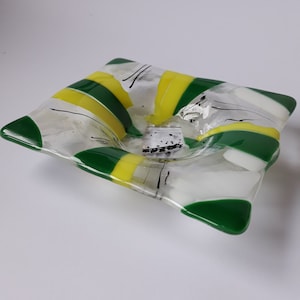 May include: A square glass dish with a white, green, and yellow striped design. The dish is slightly curved inward and has a small, shallow depression in the center.