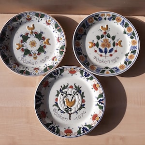 Op de afbeelding: Drie witte decoratieve borden met kleurrijke bloemen- en vogelontwerpen. Elk bord heeft het woord "Påsken" en een jaartal, 1976 of 1974. De borden hebben een geschulpte rand met blauwe, groene en oranje accenten.