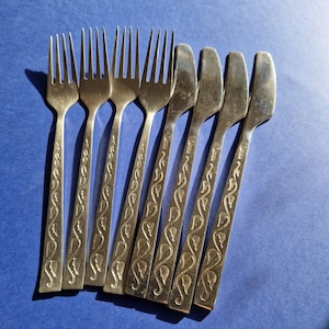May include: A collection of vintage cutlery, comprising forks and knives, displayed on a blue surface. The silver-toned cutlery features a decorative leaf pattern on the handles. The forks have four prongs, and the knives have rounded blades.