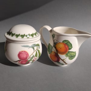 May include: A white ceramic sugar bowl and creamer set with a peach and apricot design. The lid of the sugar bowl has a green leaf border.