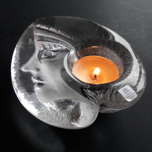 Könnte beinhalten: Ein durchsichtiger Glas-Kerzenhalter in Form eines Gesichts mit einem brennenden Teelicht darin. Der Halter ist signiert mit "Mats Jonasson Kosta Boda".