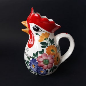 Peut inclure: Pichet en céramique blanche avec un motif de tête de coq. La crête du coq est rouge et le bec est jaune. Le pichet est décoré de fleurs colorées et de feuilles vertes. Le texte "UPP TUPP" est écrit sur le pichet.
