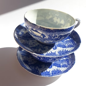 Peut inclure: Une tasse à thé et des soucoupes en porcelaine bleues et blanches. La tasse est posée sur deux soucoupes, toutes présentant un motif de paysage détaillé. Le motif est bleu sur fond blanc, typique de la porcelaine traditionnelle.