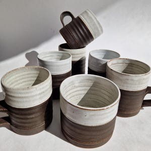 May include: A collection of handmade ceramic mugs. The mugs feature a two-tone design with a dark brown base and a white upper section. The mugs have a textured, ribbed appearance on the brown section and a smooth, glazed finish on the white section. Each mug has a handle.