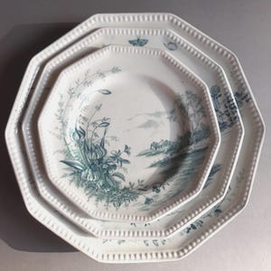May include: Three white octagonal plates with a teal floral and landscape design. The plates have a beaded edge. The largest plate is at the bottom, with the smallest plate at the top. The plates are stacked.