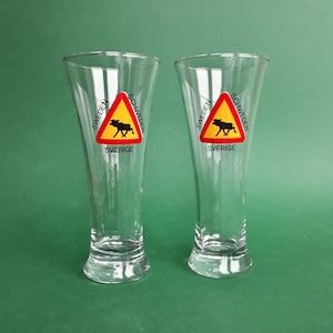 Peut inclure: Deux verres à bière transparents avec un motif de panneau routier triangulaire. Le panneau présente une silhouette de wapiti noir sur fond jaune, encadrée d'un bord rouge, avec les mots "SWEDEN" et "SVERIGE".