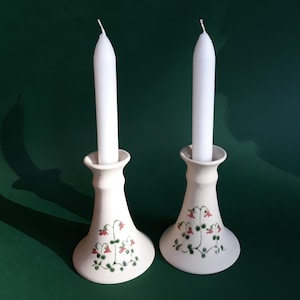 May include: Two white ceramic candle holders with white candles. The candle holders have a flared base and a floral design in red and green. The background is a dark green.
