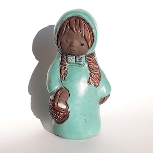 May include: A ceramic figurine of a girl in a hooded teal coat, holding a basket. The figurine has brown hair in plaits and a dark complexion. The figure is standing on a white surface.