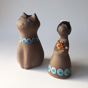May include: Two brown ceramic figurines. One is a cat with blue eyes and a blue and white collar. The other is a stylized figure of a woman with a white collar and holding dried flowers. Decorative home decor.