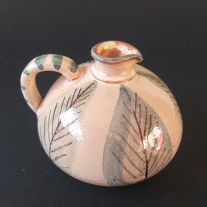 Può includere: Un piccolo boccale in ceramica dipinto a mano con una glassa rosa chiaro e un motivo a foglie verdi e marroni. Il boccale ha un corpo rotondo e un manico corto e curvo.