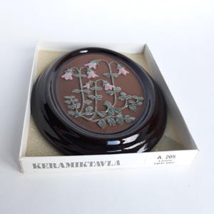 May include: A ceramic wall plaque with a brown background and a raised design of pink flowers with green leaves. The plaque is in a wooden frame and is labelled "KERAMIKTAVLA" and "A. 205 LINDA SMALAND".