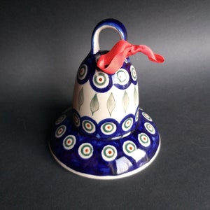Pode incluir: Um sino de cerâmica azul e branco com uma fita vermelha. O sino é decorado com um padrão de olho de pavão e folhas verdes.