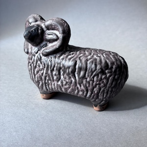 Op de afbeelding: Een keramieken ram-beeldje met een donker, getextureerd, metaalachtig glazuur. Het beeldje heeft gekrulde hoorns, een gedetailleerde wollen vacht met diepe groeven en kleine, ongeglazuurde, terracotta-kleurige voetjes aan de onderkant.