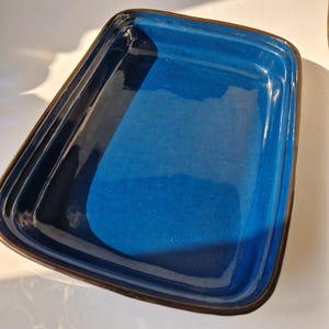 Könnte beinhalten: Eine rechteckige Backform aus Keramik mit glänzend tiefblauem Innenraum und dunklem braunen Rand. Die Form hat leicht abgerundete Ecken und eine glatte Oberfläche, geeignet für den Ofen. Das Design ist einfach und funktional.