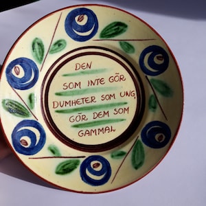 Può includere: Un piatto in ceramica giallo con un motivo floreale blu e verde. Il piatto ha una citazione in svedese che recita: "Den som inte gör dumheter som ung, gör dem som gammal."