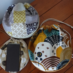 May include: Three drawstring bags with different patterns. The bags are made of fabric and have a round shape. The patterns include a patchwork design, a geometric design, and a floral design. The bags are sitting on a wooden surface.