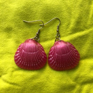 Pode incluir: Um par de brincos de concha rosa com ganchos prateados. As conchas têm um acabamento perolado e uma textura detalhada.