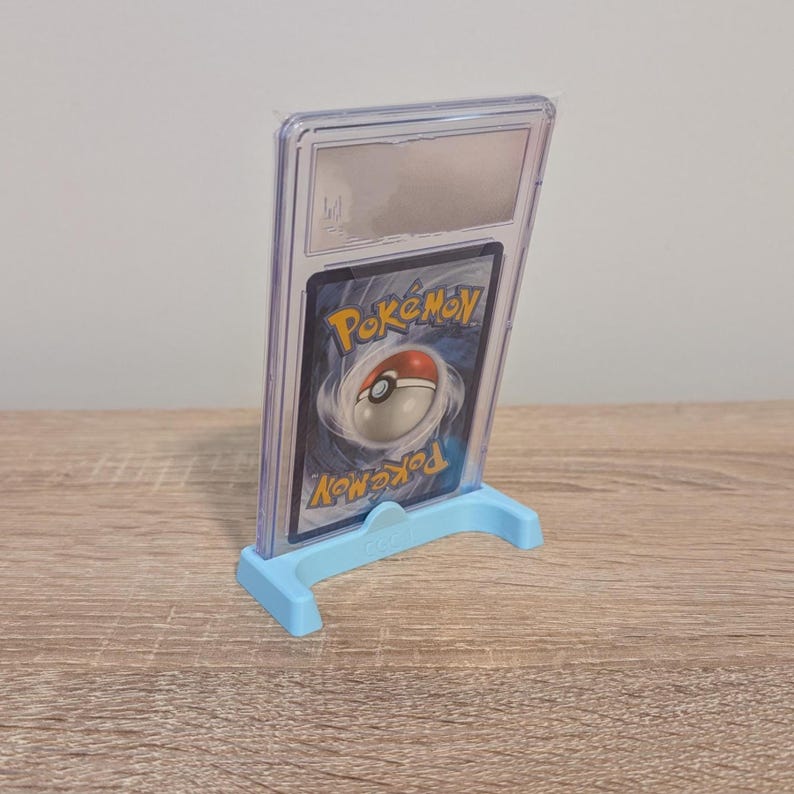 May include: A graded Pok&eacute;mon trading card encased in a clear plastic holder, displayed on a light blue stand. The card features the Pok&eacute;mon logo and a red and white Pok&eacute; Ball design. The stand is on a wooden surface.
