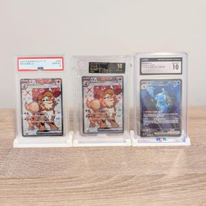 May include: Three graded Pok&eacute;mon trading cards in protective cases. The cards feature illustrations of Arcanine and Blastoise. Each case is graded a 10. The cards are displayed on white stands against a light brown surface.